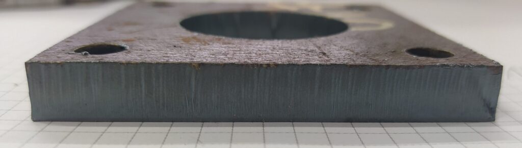 Laser-cut steel plate showing a clean, straight cut edge with visible vertical striations typical of fiber laser cutting. The surface discoloration near the edge indicates thermal influence while maintaining good dimensional accuracy.