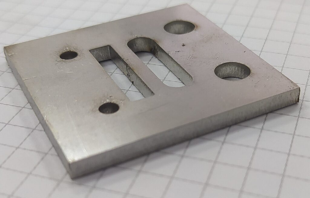 Metal plate with laser-cut holes and slots, showing clean edges and minor surface discoloration from the cutting process.