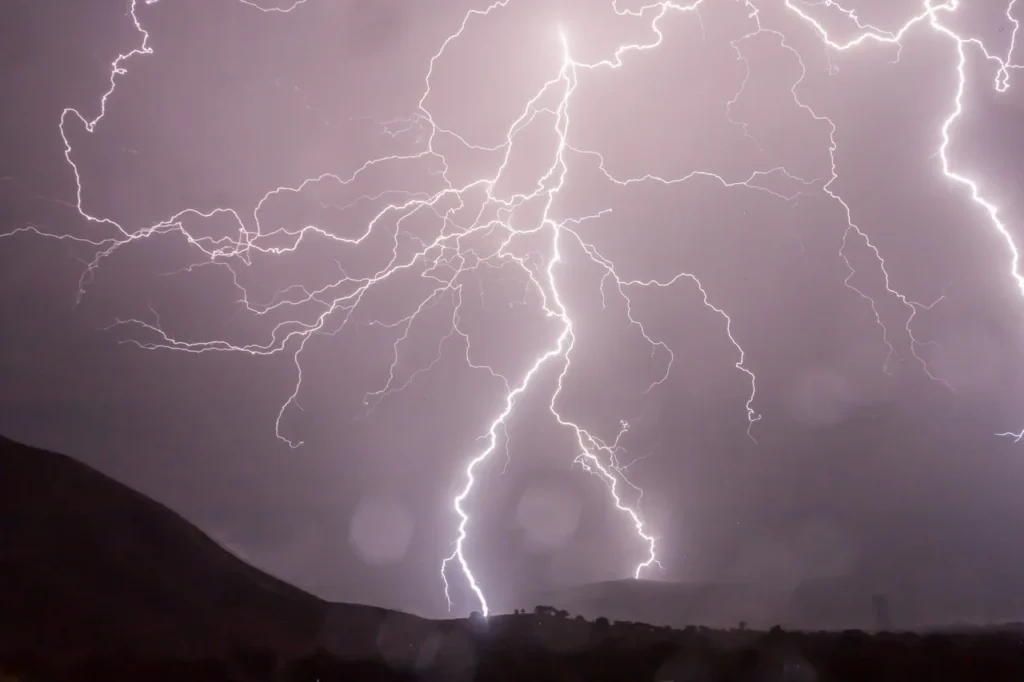 A powerful lightning strike during a thunderstorm, with multiple branching bolts illuminating the night sky above a dark, mountainous landscape—an intense display of atmospheric electrical discharge.