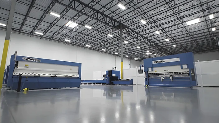 Spacious industrial showroom with Otinus CNC machines, including press brakes and a fiber laser cutting system, arranged on a polished concrete floor under a high steel-truss ceiling.